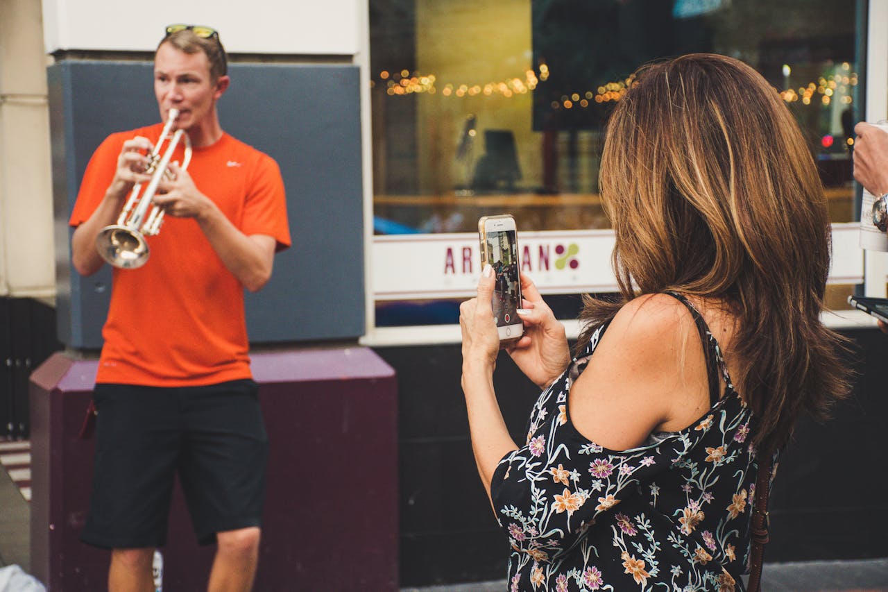 A street performer playing trumpet is photographed by a passerby in an urban setting.